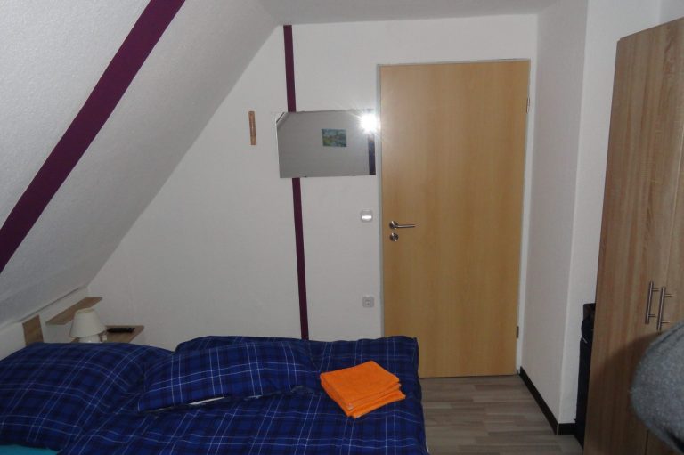 Ein kleines Zimmer mit schrägem Dach, Bett und Holzschrank neben einer Tür.